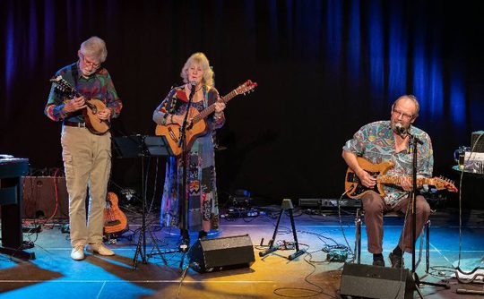 A trio of musicians performs on stage under vibrant blue lighting. The woman at the centre sings and plays guitar, flanked by a man with a mandolin and another seated with an electric guitar. The scene conveys a lively and engaging musical atmosphere.