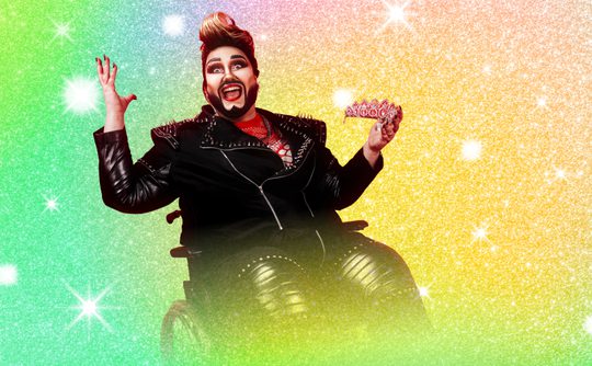Drag performer in a black studded leather outfit and wheelchair, holding a pink tiara, set against a glittering rainbow background, exudes joy and confidence.