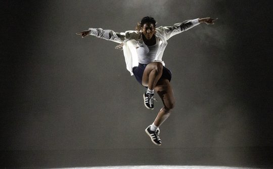 A hip-hop dancer in mid-air, arms outstretched confidently, wearing a white shirt, dark shorts, and sneakers against a dim, spotlighted background. Energetic and focused.