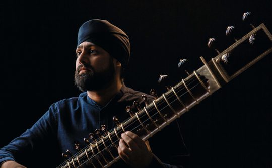 Man in a dark turban and shirt plays a sitar against a black background. His expression is focused and thoughtful, highlighting musical passion.