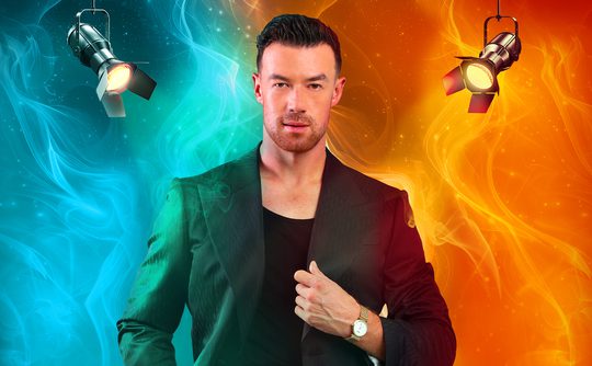 A confident man in a black suit poses against a vibrant background split into blue and orange with smoke effects. Two spotlights spotlight him from above.