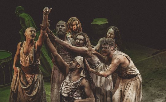 A group of performers in tattered, dusty clothing express dramatic tension on stage. They reach out, displaying intense emotions and dynamic energy.