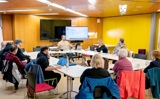 A group of people sit around a large table in a conference room, attentively listening to a speaker in front of a presentation screen. The mood is focused and collaborative.