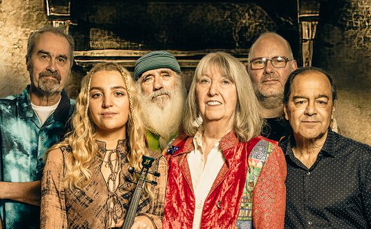 A group of six people stand close together against a textured, rustic background. One holds a violin. They appear relaxed and content, showcasing a mix of styles and ages.