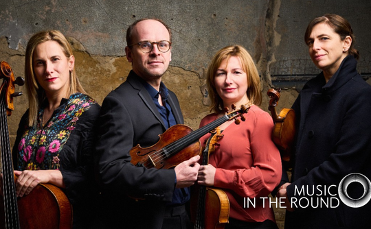 A quartet stands confidently with their string instruments against a rustic wall. They exude professionalism. Logo reads: "Music in the Round."