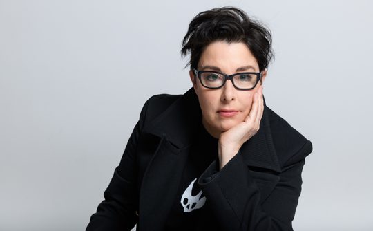 Sue Perkins, a woman with short dark hair and glasses wearing a black coat and shirt with a white logo. They rest their chin on their hand, conveying a thoughtful expression.