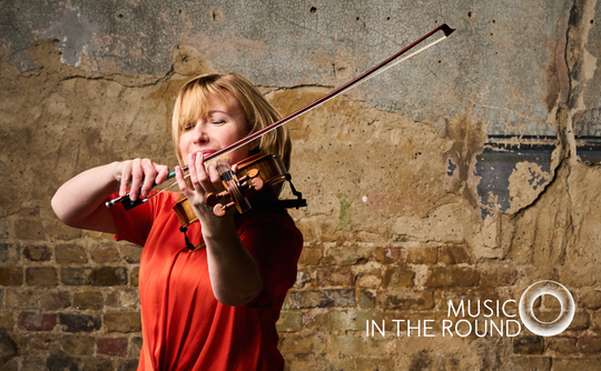 A woman in a red shirt passionately plays a violin against a rustic, textured brick wall. The logo “Music in the Round” is visible in the corner.