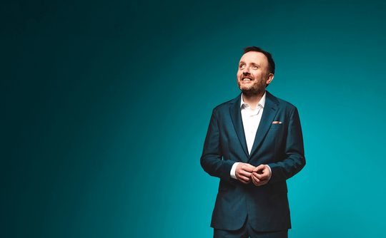 Chris McCausland, a man in a dark suit and white shirt smiles looks upward against a teal background, conveying optimism and a professional tone.