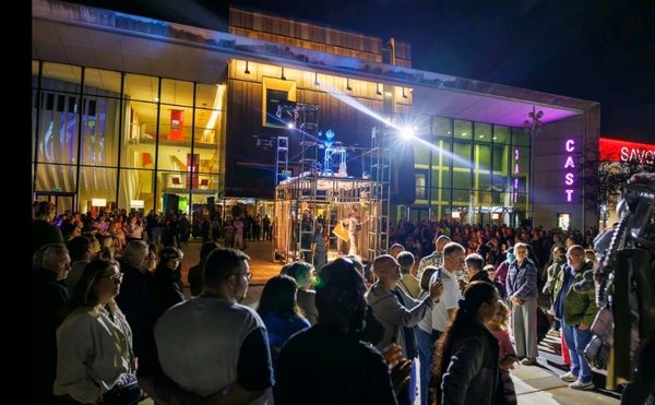 Crowd gathered at night outside a lit-up modern building, watching a street performance on a platform with scaffolding. The atmosphere is lively and engaging, taken during Lost Dream by Teatr Ósmego Dnia.