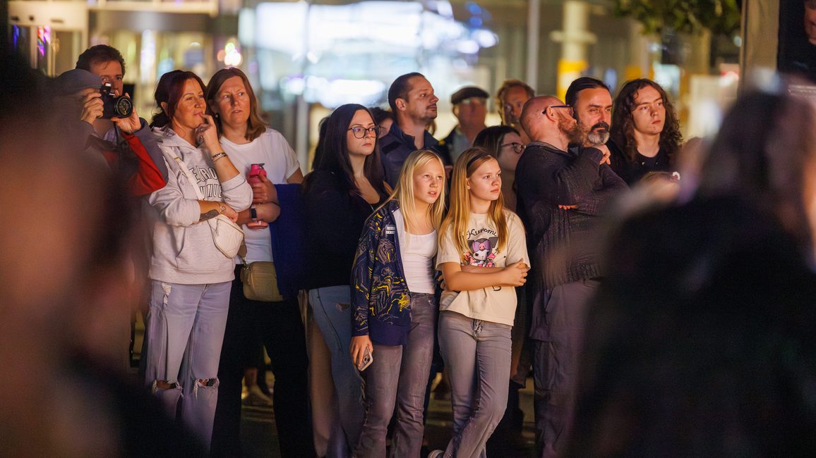 A diverse group of people stands outdoors at night, attentively watching Lost Dream by Teatr Ósmego Dnia at Cast in Doncaster. The scene is illuminated with warm light, creating an atmosphere of focus and curiosity.