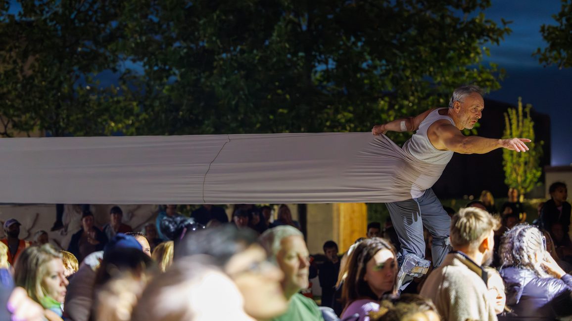 A performer balances on stilts above a crowd at night with a long fabric stretched outs behind him pulling him back as he reaches for something out of the image. There trees and the evening sky in the background. The atmosphere is tense yet captivating during a performance of Lost Dream at Cast in Doncaster by Teatr Ósmego Dnia.