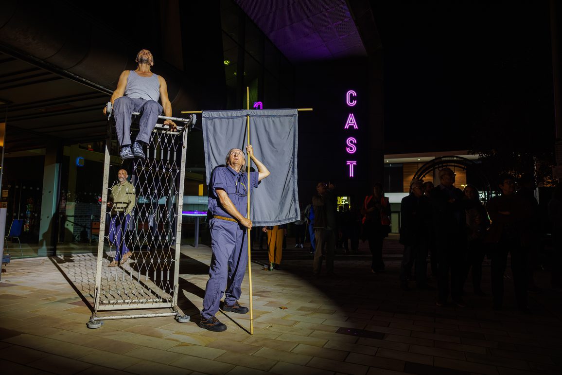 A performer sits atop a overturned metal bed frame, another stands holding a large flagpole, outside a building with neon "CAST" sign, surrounded by an evening crowd. Lost Dream by Teatr Ósmego Dnia.