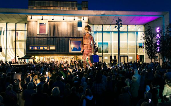 A giant puppet stands in front of the Cast building at night, the square is filled with people holding phones and taking photos