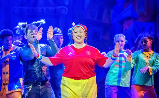 Performers on stage, brightly lit, centre joyfully gesturing. Ensemble in costumes, vibrant colours with blue backdrop. Energetic, joyful atmosphere.