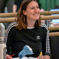 A woman in a black Adidas shirt sits smiling at a table with a notebook, pen, and snacks. She appears engaged and relaxed with a green curtain backdrop.