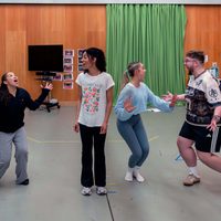 Four people are in a spacious room with wooden walls and green curtains, energetically rehearsing. They each have dynamic poses and express joy.