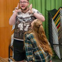 A man playfully poses with raised hands and an animated expression, while a woman in plaid looks up at him smiling. They're in a colourful, lively setting.
