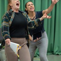 Two women passionately rehearse a scene in front of green curtains. The woman in front holds a script, wearing a plaid shirt, and they both express intense emotion.