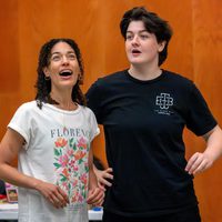 Two people stand together in a room, looking upward with expressions of excitement and curiosity. One wears a floral shirt, the other a black T-shirt.