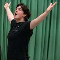 Person in a black t-shirt stands with arms raised, mouth open, and an expressive face against a green curtain, conveying excitement or joy.
