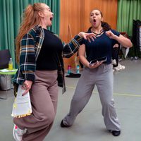 Two women energetically rehearse in a studio, with one holding papers and both expressing excitement. The room has colourful curtains and chairs.