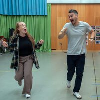 Two people are enthusiastically running indoors on a smooth floor. The woman wears a plaid shirt, and the man is in a grey t-shirt. Both look joyful and animated, set against a background of colourful curtains and wooden panels.