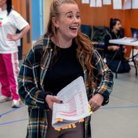 A woman with long hair, wearing a checked shirt, smiles while holding a script with colourful tabs in a rehearsal room. Joyful, focused atmosphere.