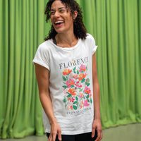 Woman wearing a floral "Florence" T-shirt laughs joyfully against a soft green curtain backdrop. The scene conveys a sense of happiness and warmth.
