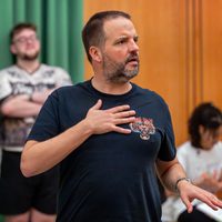 Man in a navy shirt with a tiger emblem gestures passionately in a room. Behind him, two blurred figures are engaged, one standing with arms crossed.