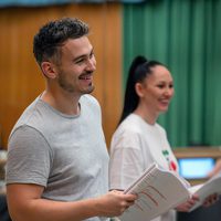 A man and a woman smiling while holding scripts in a bright rehearsal space, suggesting a joyful, collaborative atmosphere. Green curtains in the background.