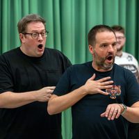 Two men express surprise with open mouths and wide eyes. One touches his chest while the other points. A green curtain serves as the backdrop.