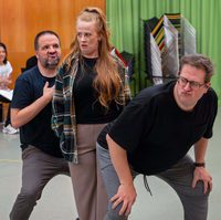 Three people in a rehearsal space make exaggerated expressions and poses. The setting is energetic and humorous, with a green curtain and chairs in the background.
