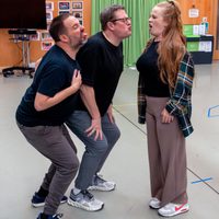 Three people humorously mimic a standoff, sticking out their tongues in a playful manner. They stand in a room with green curtains and wooden walls.