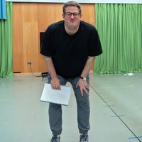Man in black shirt and glasses, leaning forward with a smile, holds a notebook. Behind him are green curtains and a wooden wall, conveying a casual setting.