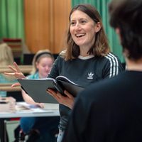 A woman with long brown hair, smiling, holds an open script in a rehearsal room with a green curtain. She wears a black shirt; others are blurred in the background.