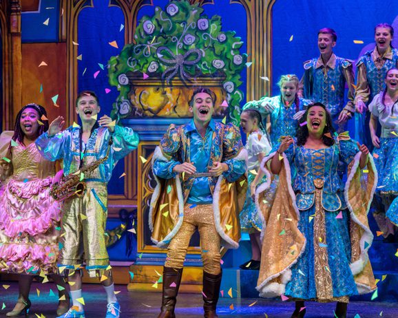A jubilant group of performers in colorful, ornate costumes sing joyously on stage amidst falling confetti. The background is a vibrant, theatrical set.