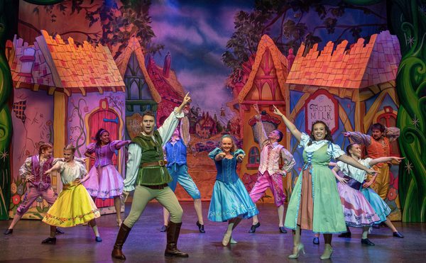 A colourful stage performance with actors in medieval attire, vibrant dresses, and fantasy scenery of cottages. The scene is lively and joyful.