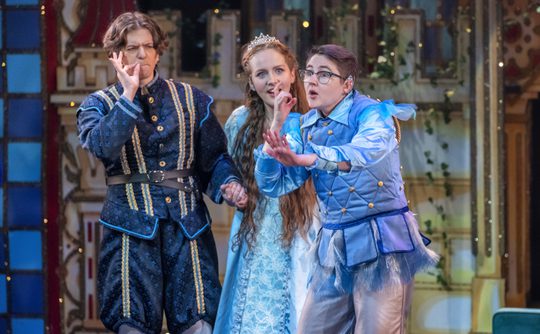 Three actors in Renaissance costumes perform on stage. The centre actor, wearing a crown, appears surprised, while the others make expressive gestures.
