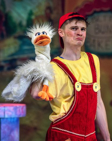 A person in a red cap and overalls holds a fluffy duck puppet, making a comical face. The colorful background suggests a playful, theatrical setting.