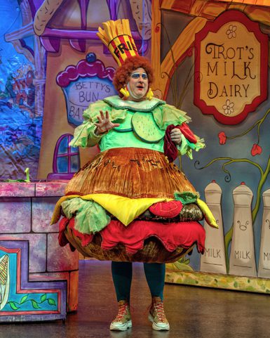 A performer in a vibrant burger costume, complete with bun, lettuce, and tomato, gestures expressively on a colorful stage with playful, whimsical set design.