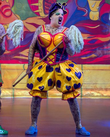 A performer in a vibrant yellow costume with blue hearts and a red bodice, holding mops. The background is colorful and abstract; the expression is dramatic.