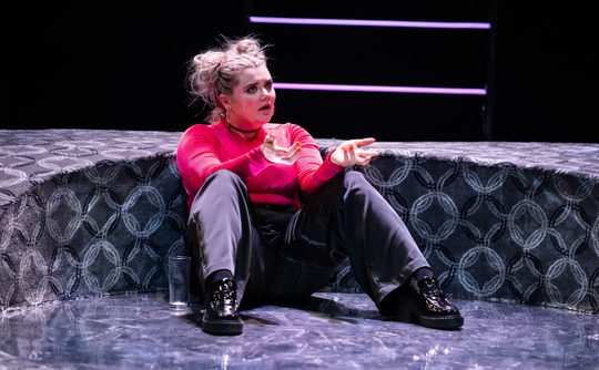 A person with a distressed expression, wearing a pink shirt and dark trousers, sits on a patterned stage floor. Their hands gesture expressively.