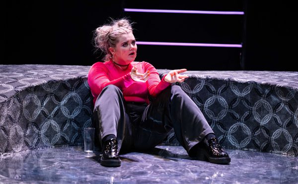 A person with a distressed expression, wearing a pink shirt and dark trousers, sits on a patterned stage floor. Their hands gesture expressively.