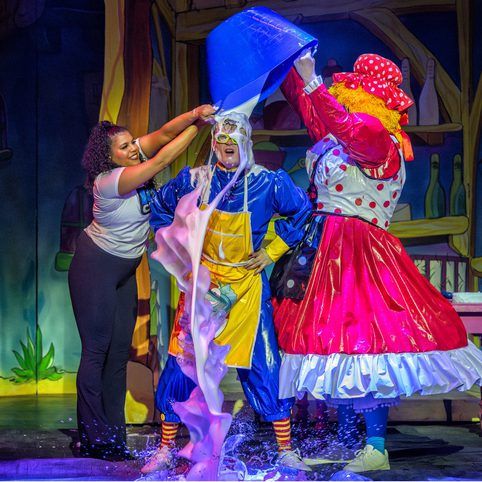 Three actors stand on stage, two actors pour sludge over the actor in the middle. The actor to the left wears a Doncaster Dome Icecaps shirt, the one on the right wears a bright red Dame outfit and the one in the middle wears blue overalls and a yellow apron.