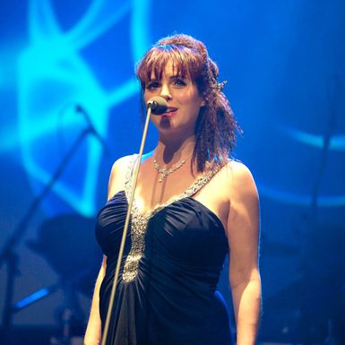 A White female performer with brown hair wearing a black dress sings into a microphone.