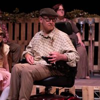 Actors on stage, three in the background sat on a set made from pallets, they all are looking to the right. Actor in the foreground is in a wheelchair and looking off to the right.