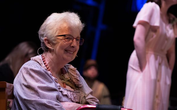 A older female actor sits looking off to the left, she is smiling and sits with her arms folded.