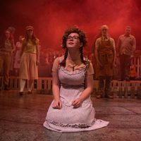 A group of actors stand bind a actor in the foreground looking on. They are cast in a red soft light and watching the female actor at the front. She looks up and slightly to the left she wears a crown of heather.