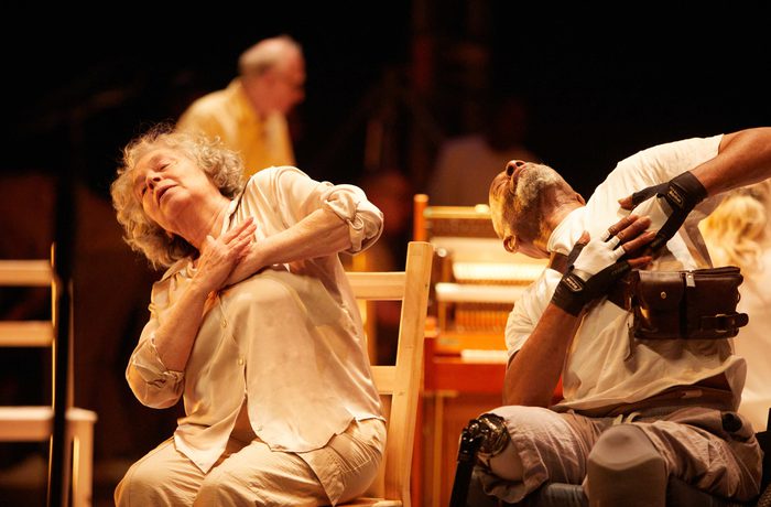 Two older performers lean back with hands over their hearts, one white woman on a wooden chair and one black man in a wheelchair.
