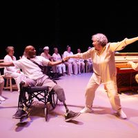 Two older performers dressed in white dance together, one White woman with grey hair and one bald Black man in a wheelchair with two prosthetic legs. A performer plays piano in the background.